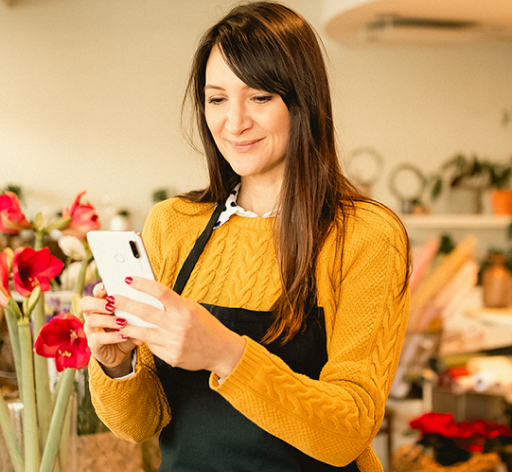 Person in yellow sweater and black apron smirking, holding a phone, standing by red flowers in a bright room.